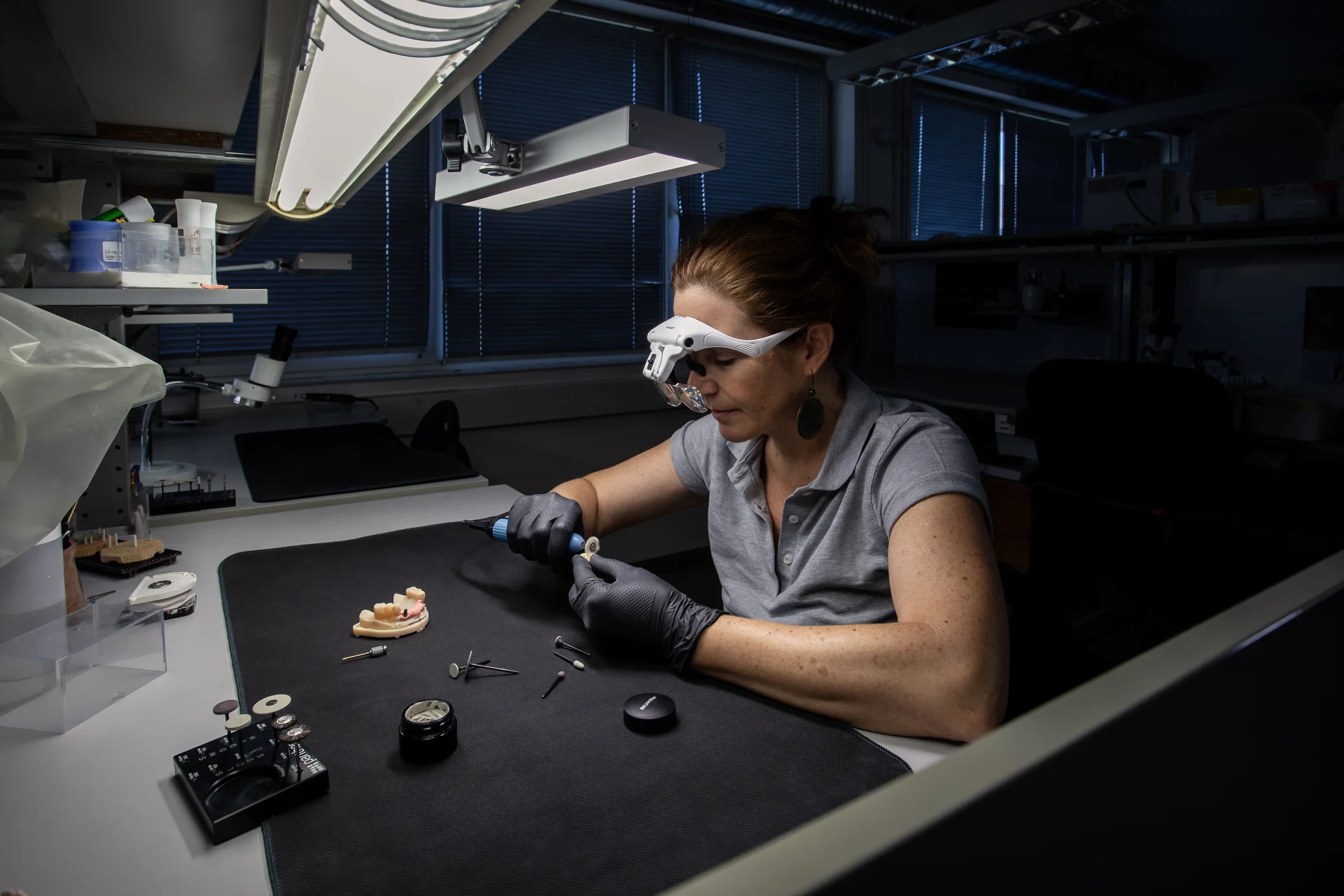 Eine Zahntechnikerin bearbeitet Zahnersatz mit Lupenbrille und Handstück im Bridge Dental Labor.